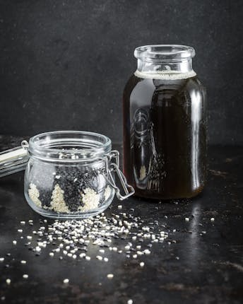 Ein Glas mit dunklem, gerösteten Sesamöl neben einem Einmachglas voll schwarzem und weißem Sesam vor dunklem Hintergrund