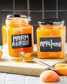 Zwei eckige Schraubgläser sind mit orangener Aprikosenmarmelade gefüllt undauf einem Holzbrett neben halbierten Aprikosen platziert.
