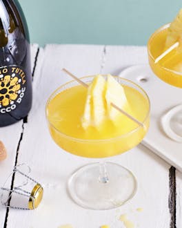 Zwei Gläser mit sonnengelbem Flirtini und Ananas-Stücken am Spieß auf einem weißen Holztisch vor türkisem Hintergrund