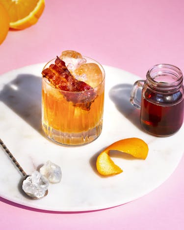Old Fashioned Cocktail mit Bacon auf einem Marmortablett mit Orangen und einem Löffel mit Kandiszucker.