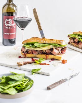 Zwei Steak & Cheese Sandwiches neben zwei gefüllten Weingläsern und einer Flasche Cabernet Sauvignon vor hellem Hintergrund