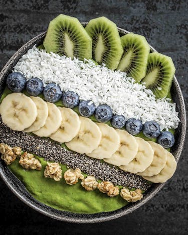 Grüne Smoothie-Bowl mit Topping-Streifen aus Kiwi, Kokosraspel, Heidelbeeren, Banane, Chia und Granola auf dunklem Untergrund