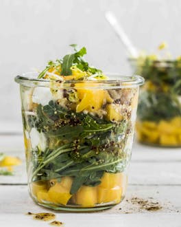 Mango-Salat mit Rucola im Glas mit Besteck auf weißem Holzuntergrund.