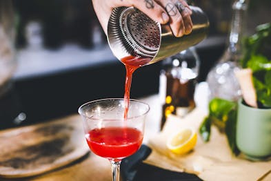 Eine Hand füllt den Jack Rose Cocktail mit einem Strainer aus dem Shaker in ein Cocktailglas.