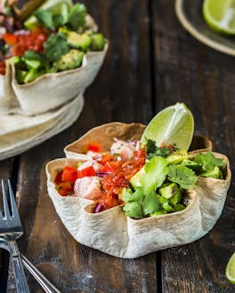 Zwei Bowls aus Tortilla-Fladen gefüllt mit Lachs, Salsa, Avocado und Koriander neben Limettenvierteln auf Holzgrund