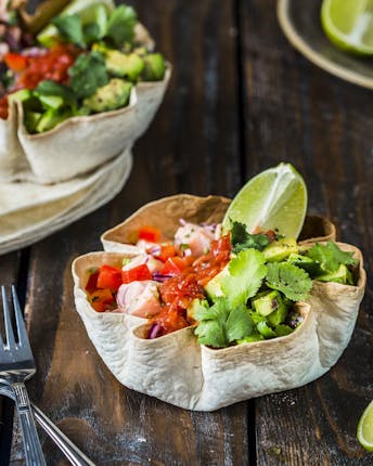 Zwei Bowls aus Tortilla-Fladen gefüllt mit Lachs, Salsa, Avocado und Koriander neben Limettenvierteln auf Holzgrund