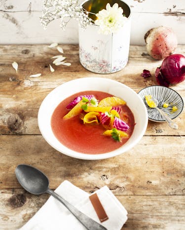 In einem weißen tiefen Teller ist eine rote Bete Suppe mit Orangenfilets. Im Hintergrund eine weiße Blume, eine Zwiebel, eine kleine Schale mit einem Teelöffel.