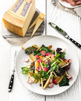 Ein weißer Teller, der mit einem Salat aus Grünkohl, Gruyère-Käsestreifen, Radieschen und Mettwurst gefüllt ist, stehen neben einer Schale mit dünn geschnittener Mettwurst und einem Stück Gruyère auf einem weiß gebeizten Holzuntergrund