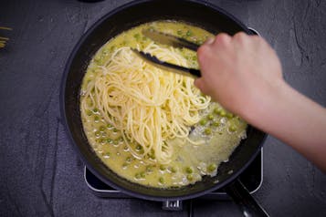 Spaghetti werden mit einer Zange mit der Erbsen-Weißweinsoße in der Pfanne vermischt.