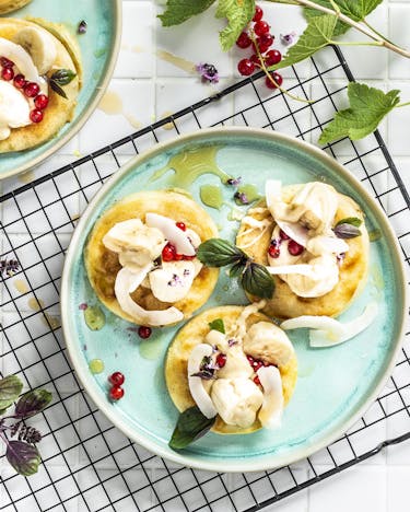 Kleine, runde Pfannkuchen auf Tellern mit Bananenscheiben, Kokoschips und Johannisbeeren