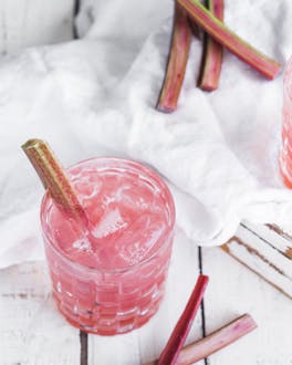 Zwei Gläser sind mit dem pastellig-pinken Drink gefüllt und mit jeweils einer Stange frischem Rhabarber dekoriert.