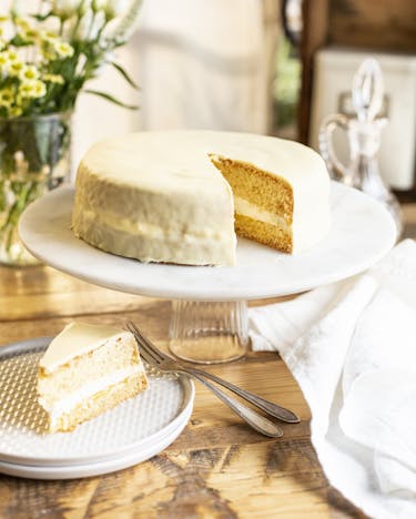 Auf einer Tortenplatte liegt eine kleine Lemon-Curd-Torte mit einer weißen Schicht zwischen den Böden. Davor ein Teller mit einem Stück der Torte