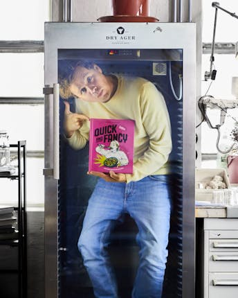 Hannes steht in einem durchsichtigen Kühlschrank und hält sein neues Kochbuch in den Händen.