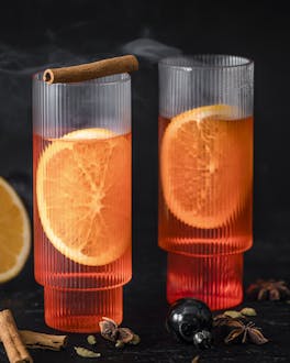 zwei Gläser mit heißem Aperol Spritz, dahinter Orangen und Zimtstangen