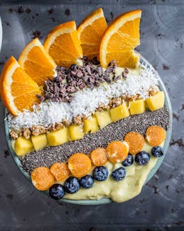 Mango-Smoothie-Bowl mit Toppings in Streifen aus Orangenspalten, Kakaonibs, Kokosraspel, Mango, Chiasamen, Physalis und Heidelbeeren auf dunklem Grund