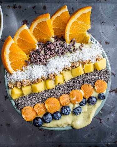 Mango-Smoothie-Bowl mit Toppings in Streifen aus Orangenspalten, Kakaonibs, Kokosraspel, Mango, Chiasamen, Physalis und Heidelbeeren auf dunklem Grund