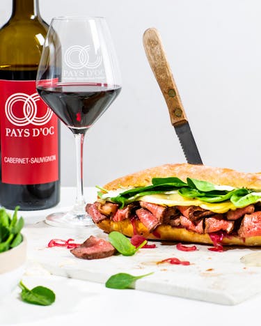 Zwei Steak & Cheese Sandwiches neben zwei gefüllten Weingläsern und einer Flasche Cabernet Sauvignon vor hellem Hintergrund