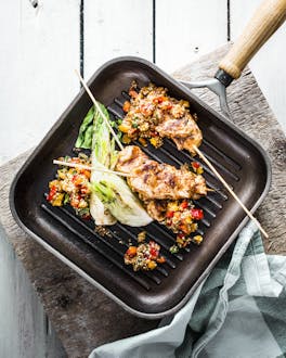In einer Grillpfanne ist ein Quinoasalat mit Hähnchen-Saté-Spießen und Pak Choi angerichtet. Dahinter ein hellgrauer Holzboden