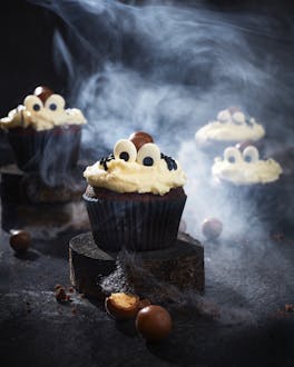 Vier Schokoladenmuffins für Halloween mit hellem Frischkäse-Frosting und einer "Spinne" aus Schokopralinen, Zuckeraugen und schwarzer Zuckerschrift für die Beine