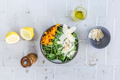 Ein Salat aus Spinat, Aprikosenfilets und Parmesan auf weißem Hintergrund. Daneben eine aufgeschnittene Zitrone, eine Pfeffermühle, ein Glasgefäß mit Öl und eine kleine graue Schale mit gehobeltem Parmesan