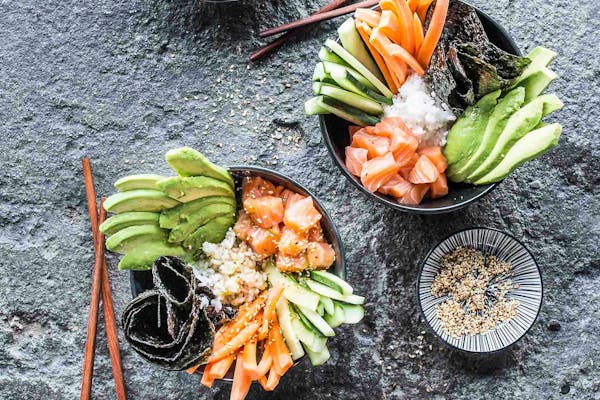 Zwei Sushibowls mit Karotten- und Gurkensticks, Avocadoscheiben, Reis, Lachsstücken und schwarzen Algen neben Essstäbchen auf grauem Stein