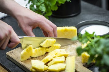 Auf einem Holzbrett wird eine geschälte Ananas in grobe Stücke geschnitten.