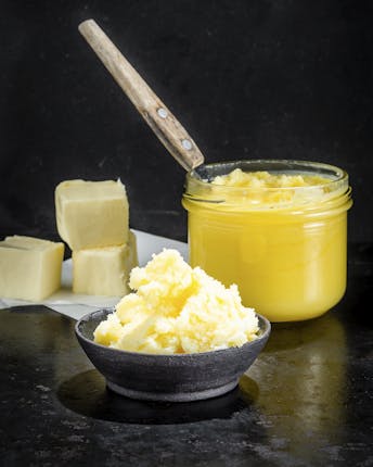 Eine Schale mit abgeschabtem Butterschmalz bzw. Ghee, ein Glas mit flüssigem Butterschmalz und ein paar feste Schmalzblöcke vor dunklem Hintergrund