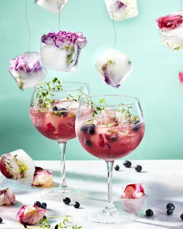 In zwei Weingläsern Blueberry Gin Spritz mit Eiswürfeln, und Blaubeeren. Um Die Drinks herum und in der Luft hängen Eiswürfel mit Blüten.