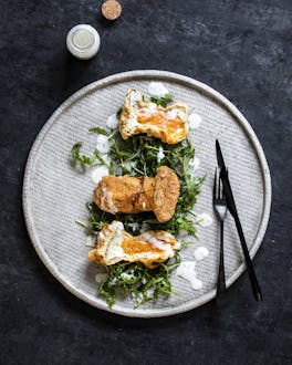 Auf einem hellgrauen Teller liegen längs aufgeschnittene, frittierte Pilze mit Käsefüllung auf einem Salatbett. Darüber ist helles Dressing