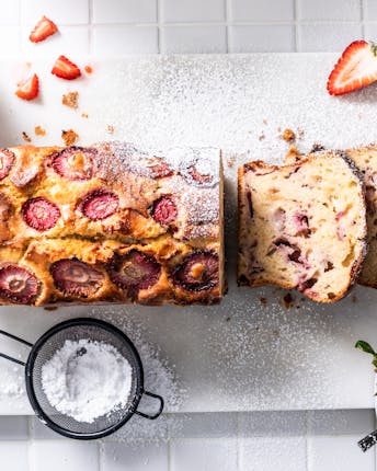 Kastenförmiges Strawberry Bread, das bereits angeschnitten ist, im Topshot von oben mit frischen Erdbeeren und einem kleinen Sieb mit Puderzucker vor hellem Fliesenhintergrund.