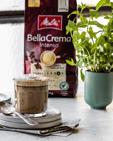 Bitterschokoladen-Cremaccino im Tumbler vor Fenster mit Melitta Bellacream Intenso Packung.
