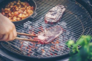 Auf einem großen Grill werden zwei Scheiben Ribeye-Steak gegrillt. Eine Hand mit Holzzange wendet eins der Steaks. Danebenm eine Pfanne mit Tomaten-Kartoffel-Würfeln.