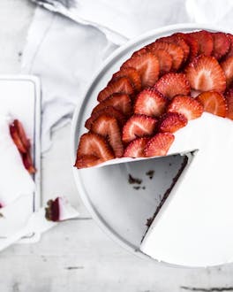 Aus einer weißen Torte, die zur Hälfte mit Erdbeerscheiben belegt ist, ist ein stück herausgeschnitten und liegt daneben auf einem weißen Brettchen,