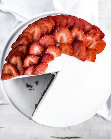 Aus einer weißen Torte, die zur Hälfte mit Erdbeerscheiben belegt ist, ist ein stück herausgeschnitten und liegt daneben auf einem weißen Brettchen,