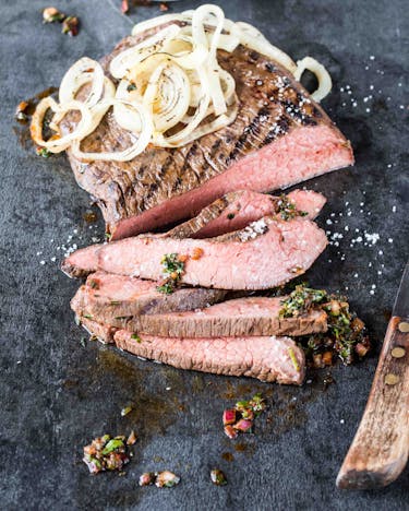 In Scheiben geschnittenes Flank Steak mit Grillstreifen und Zwiebeln auf grauem Untergrund.