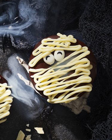 Mehreremit Schokolade glasierte Donuts, die für Halloween wie Mumien mit weißer Schoko-Ganache und Zuckeraugen dekoriert sind