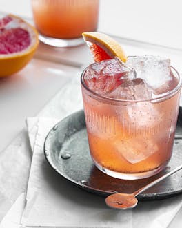 Ein Glas mit rot-oranger Rhabarber-Grapefruit-Limonade und eine halbierte Grapefruit.