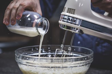 Aus einer Glasflasche wird Milch in eine Glasschüssel gegeben und mit einem Mixer verrührt.