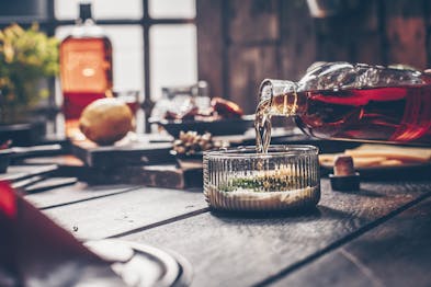 Whiskey wird in eine Glasschüssel mit Mayonnaise und Gewürzen gekippt, unscharf im Hintergrund eine Flasche Whiskey, eine Zitrone und Geschirr und Bretter