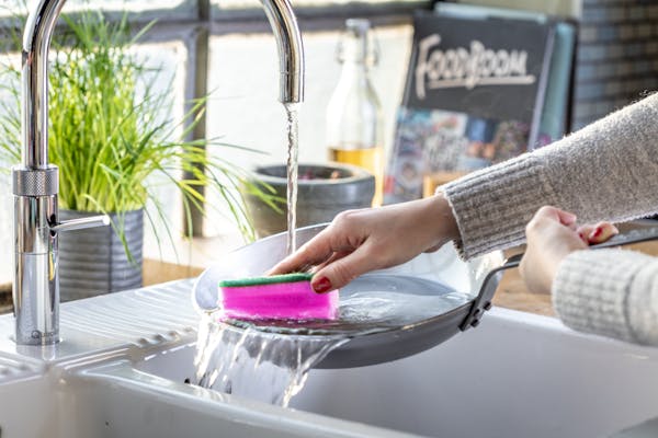 Eine Bratpfanne wird im Emaille-Spülbecken von einer weiblichen Hand mit einem pinken Schwamm und fließendem Wasser gereinigt