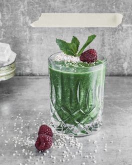 Glas mit Grünkohl-Smoothie getoppt mit Himbeere, Minze und Amaranth vor grauem Grund