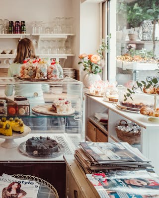 Quelle: Pay Now Eat Later. Café und Confiserie Hosch in Hamburg – die Auslage von vielen Kuchen und Törtchen ist zu sehen, eine Mitarbeiterin agiert im Hintergrund.