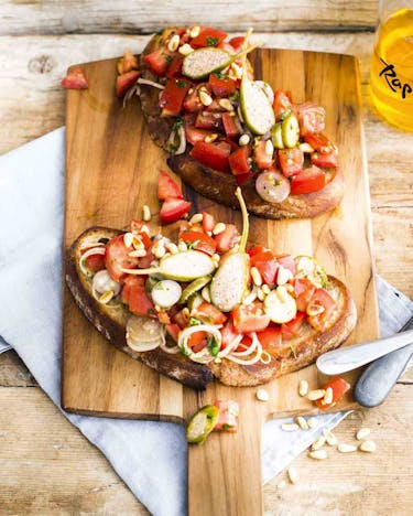 Zwei Scheiben Brot mit Tomaten-Kapernsalat auf kleinem Holzbrett mit angelegtem Besteck.
