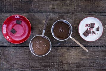 Auf einem Holzuntergrund stehen zwei Töpfe mit geschmolzener Schokoladen, daneben zwei Teller mit Kuvertüre-Stücken