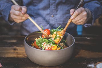In einer großen grauen Schüssel wird ein Salat mit Tomaten, Aprikosen, Blattspinat und Kichererbsen wird mit zwei Holzlöffeln vermengt.