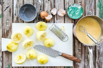 Halbierte, abgeriebene und ausgepresste Zitronen auf einem weißen Brett. Daneben eine kleine Reibe und ein großes Messer. Außerdem ein Topf mit gelber Flüssigkeit