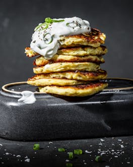 Ein Stapel von goldbraun gebackenen Kartoffel-Pancakes mit einem Klecks Crème fraîche on top und Frühlingszwiebeln vor dunklem Hintergrund