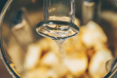Ein Flaschenhals aus Glas im Fokus, aus dem Flüssigkeit in einen unscharfen Mixer mit gelben Früchten gegossen wird.