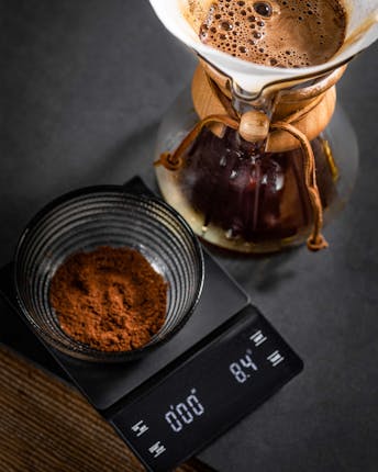 Feines Kaffeepulver, das in einer Glasschüssel auf einer Küchenwaage abgewogen wird, steht neben einer Chemex mit Handfliter, durch den frischer Kaffee läuft