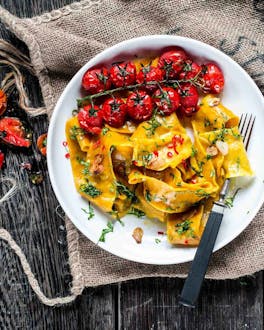 Breite Pasta aglio e olio mit geschmorten Tomaten auf einem weißen Teller, fotografiert auf einem alten Jutesack und auf dunklem Holzuntergrund.